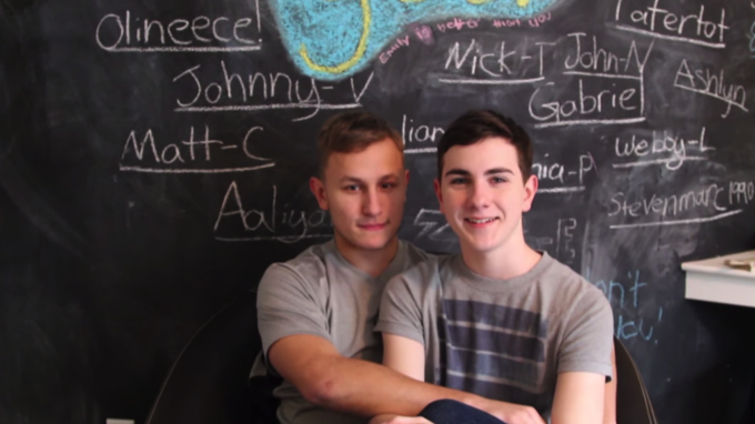 Screenshot of Austin Wallace's video, in which he is being hugged by another boy in front of a school chalkboard while he explains why he had to leave his private school. 